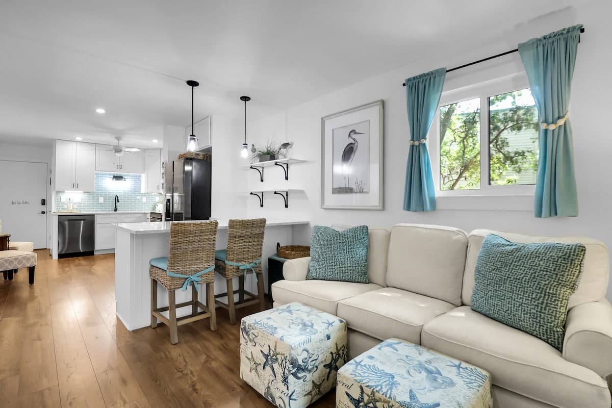 Open-concept living room with coastal-chic sofa, wicker bar stools, and kitchen